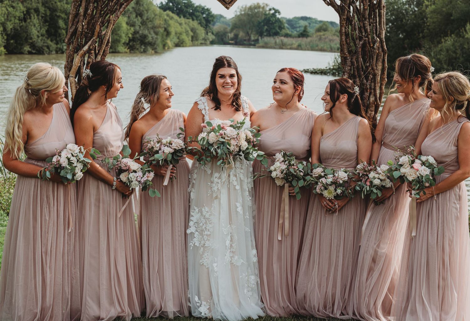 Bridal party in flowing blush pink dresses stands together by a lake holding neutral floral bouquets.