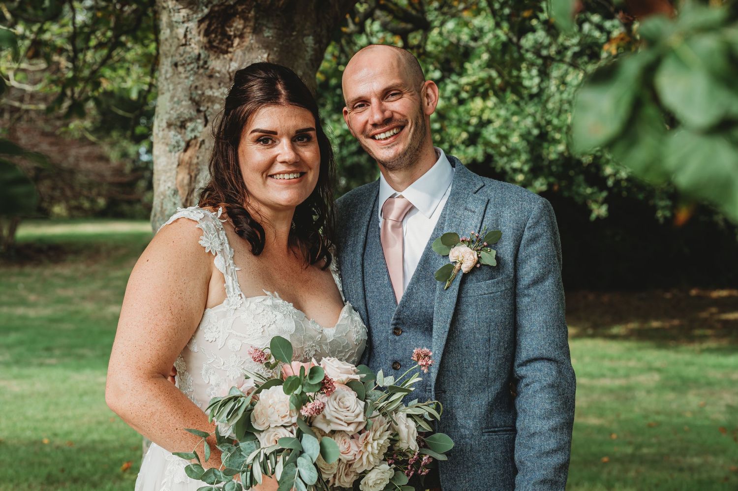 A couple in wedding attire poses outdoors on a sunny day with a bouquet and greenery in the background.
