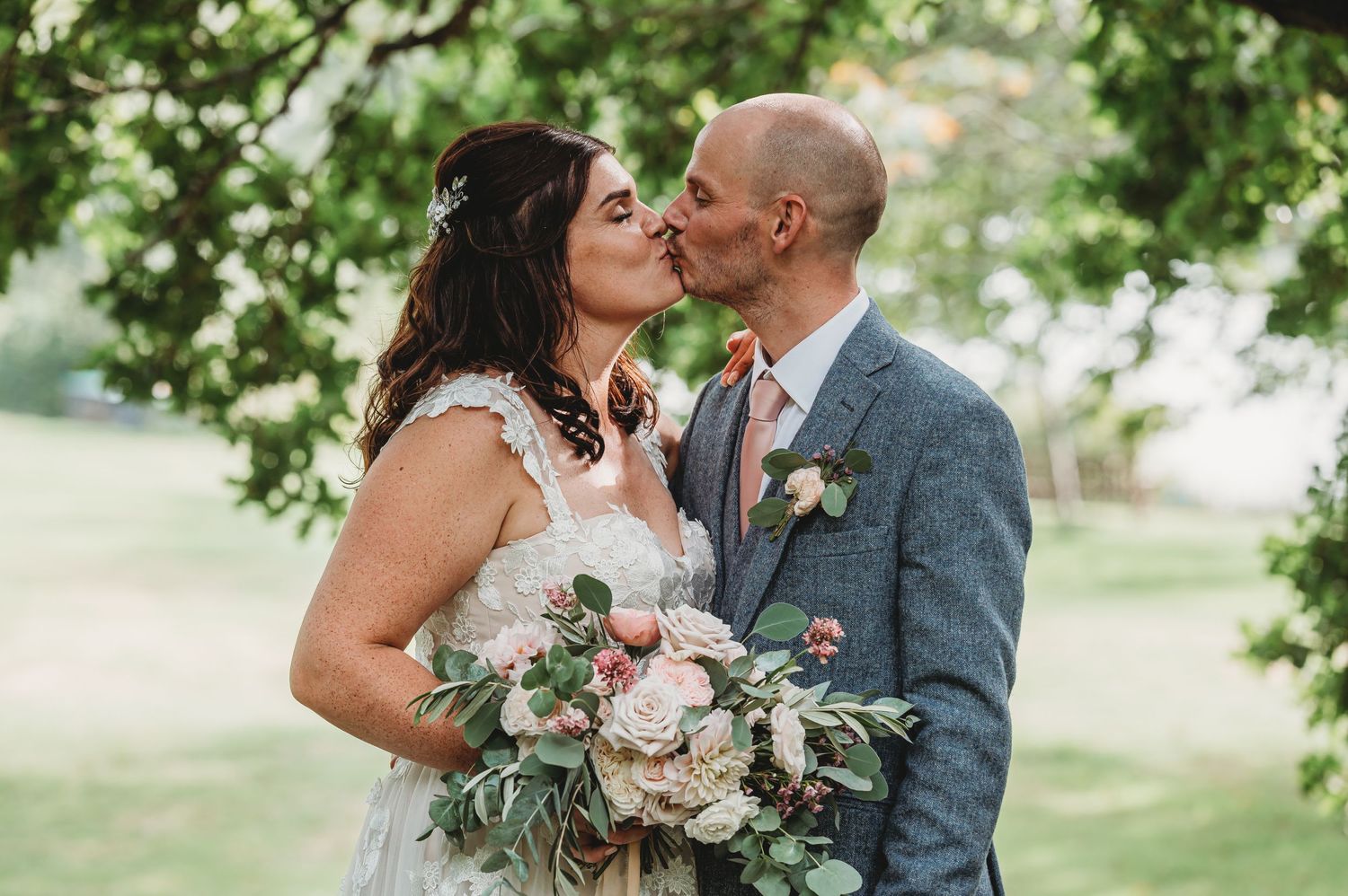 Romantic wedding couple shares a kiss outdoors under trees while holding a blush and eucalyptus bouquet.