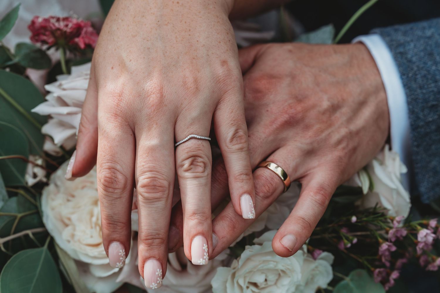 Two hands wearing wedding rings rest together on white roses and floral decorations.