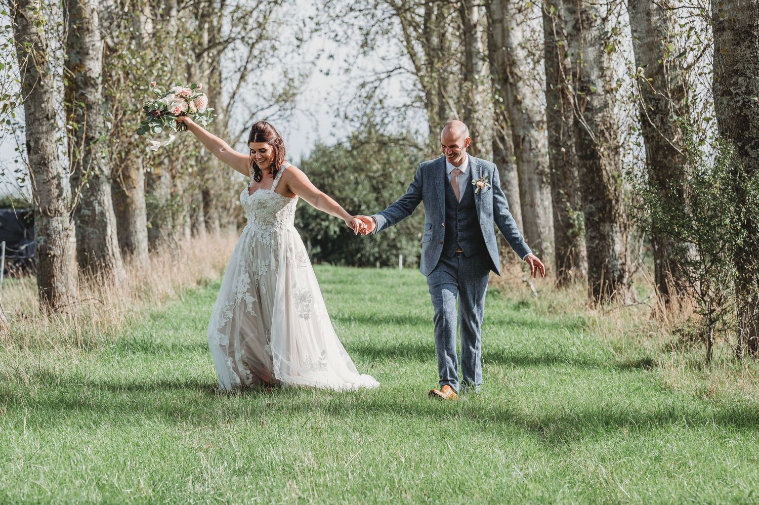 A joyful couple runs hand-in-hand through a tree-lined path on their wedding day in an outdoor setting.