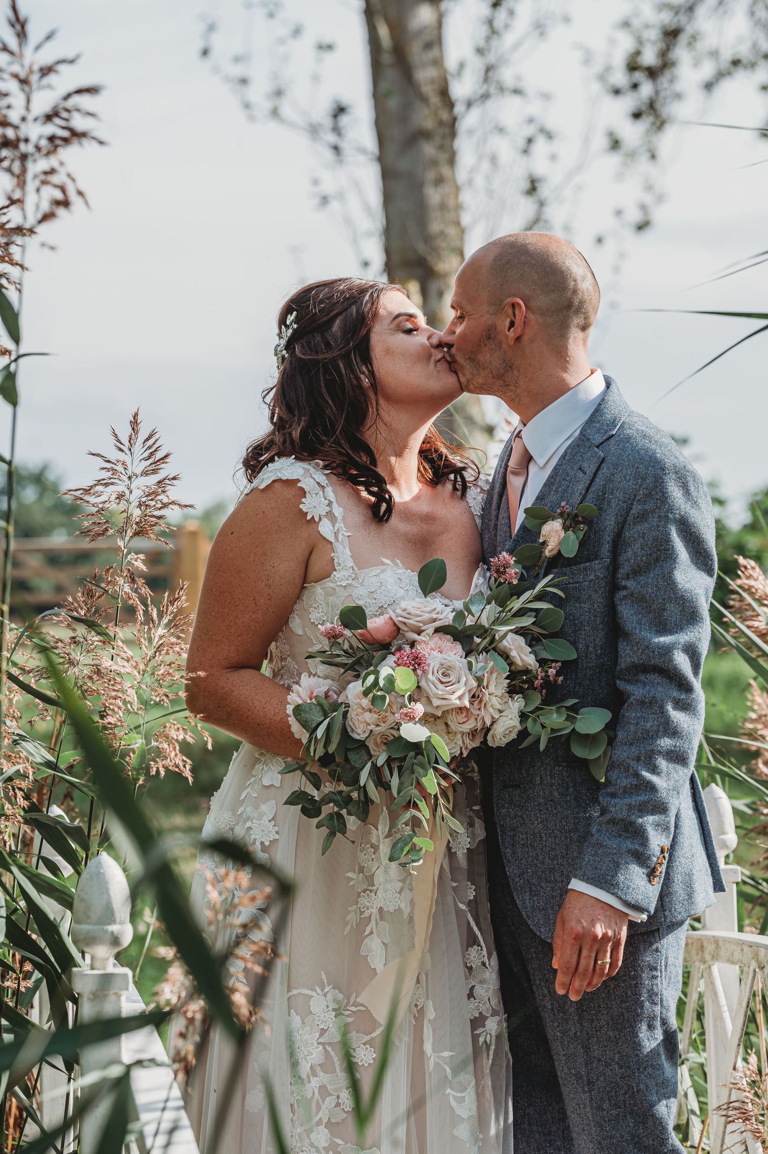 A couple shares a romantic kiss at their outdoor wedding ceremony while holding a beautiful blush and cream bouquet.