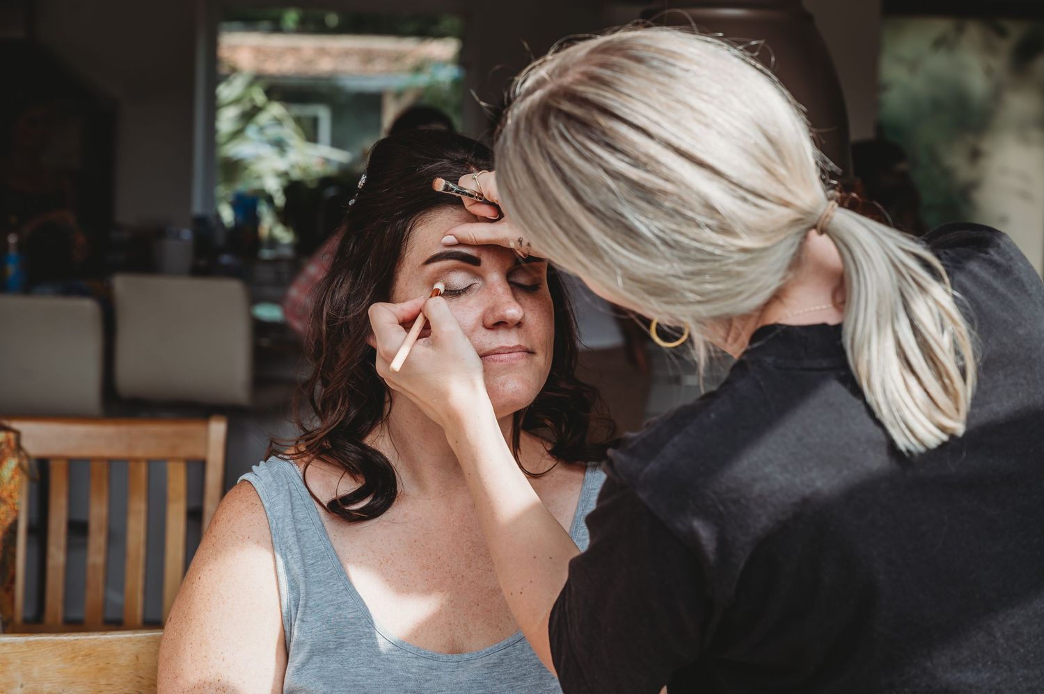 Makeup artist in black applies cosmetics to client in gray tank top during beauty session.