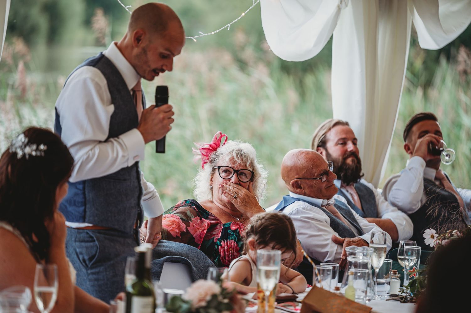 A wedding reception guest gives a speech while others at the table listen during an outdoor celebration.