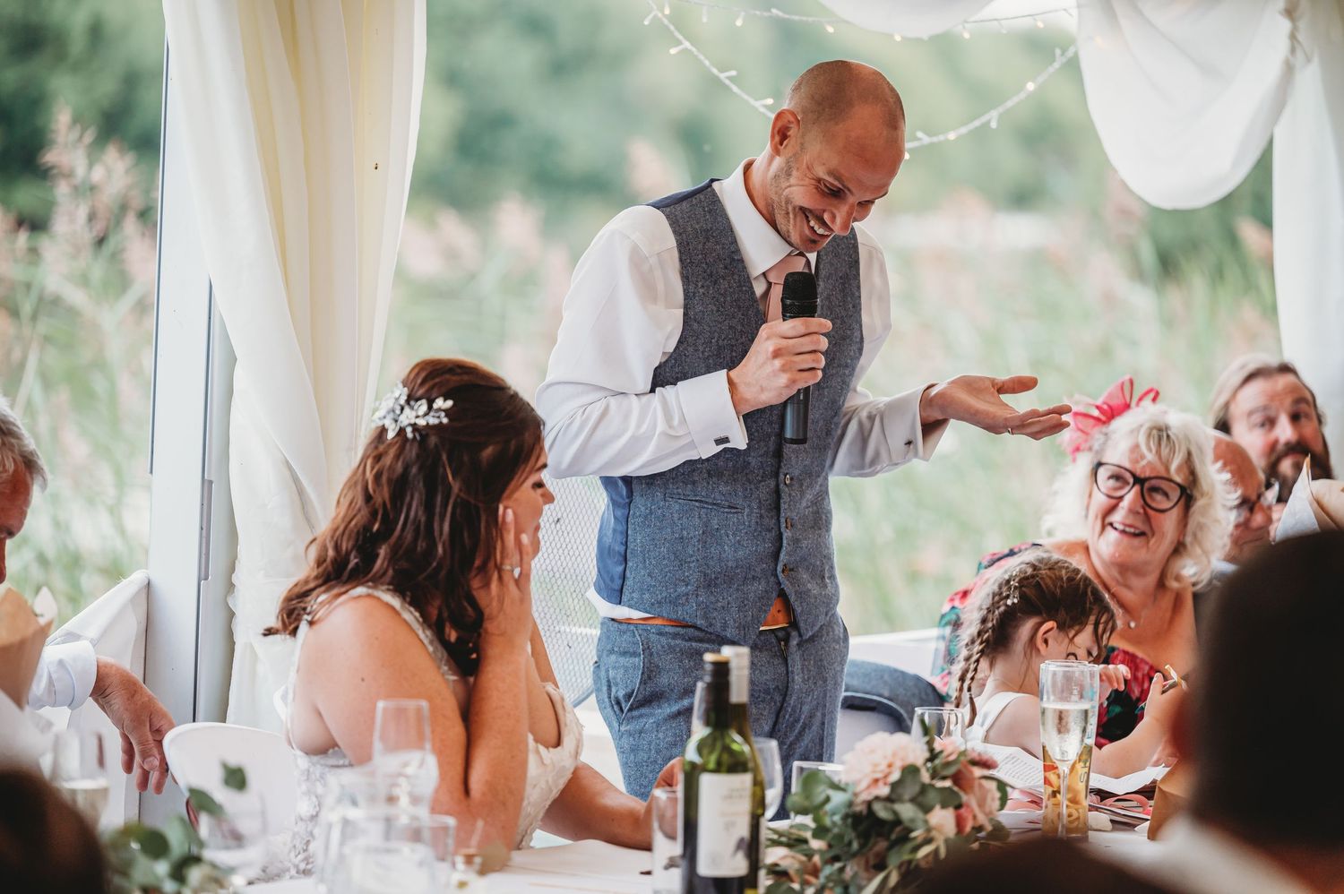A joyful wedding reception moment under a white tent as a guest in a vest delivers a speech at the head table.