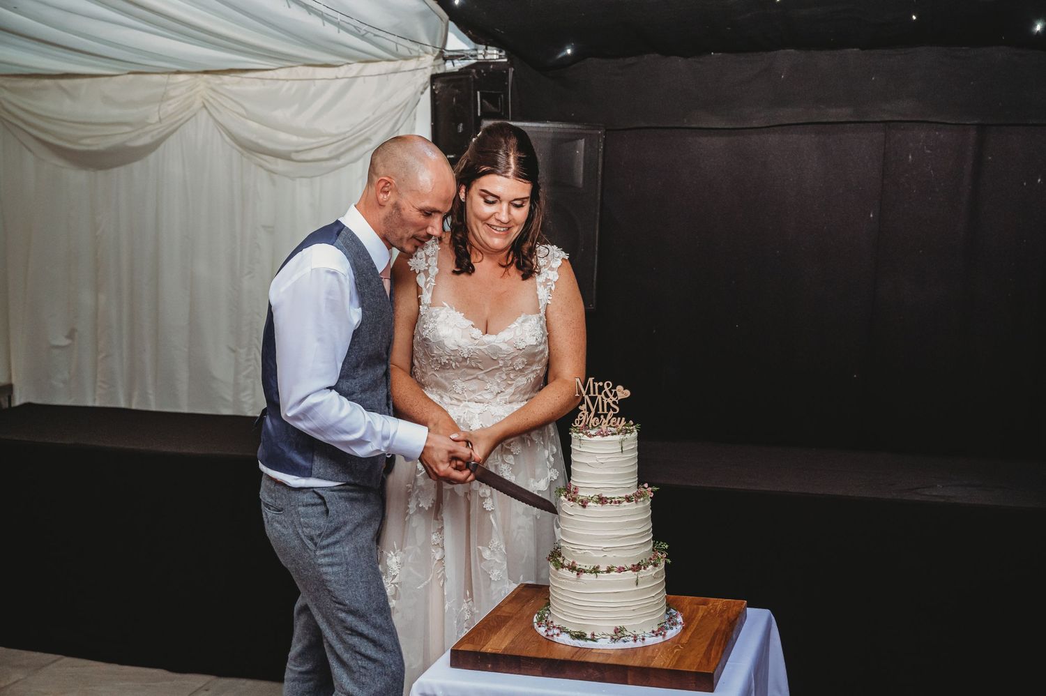 A couple in formal wedding attire stands together at their reception cutting their three-tiered white wedding cake.