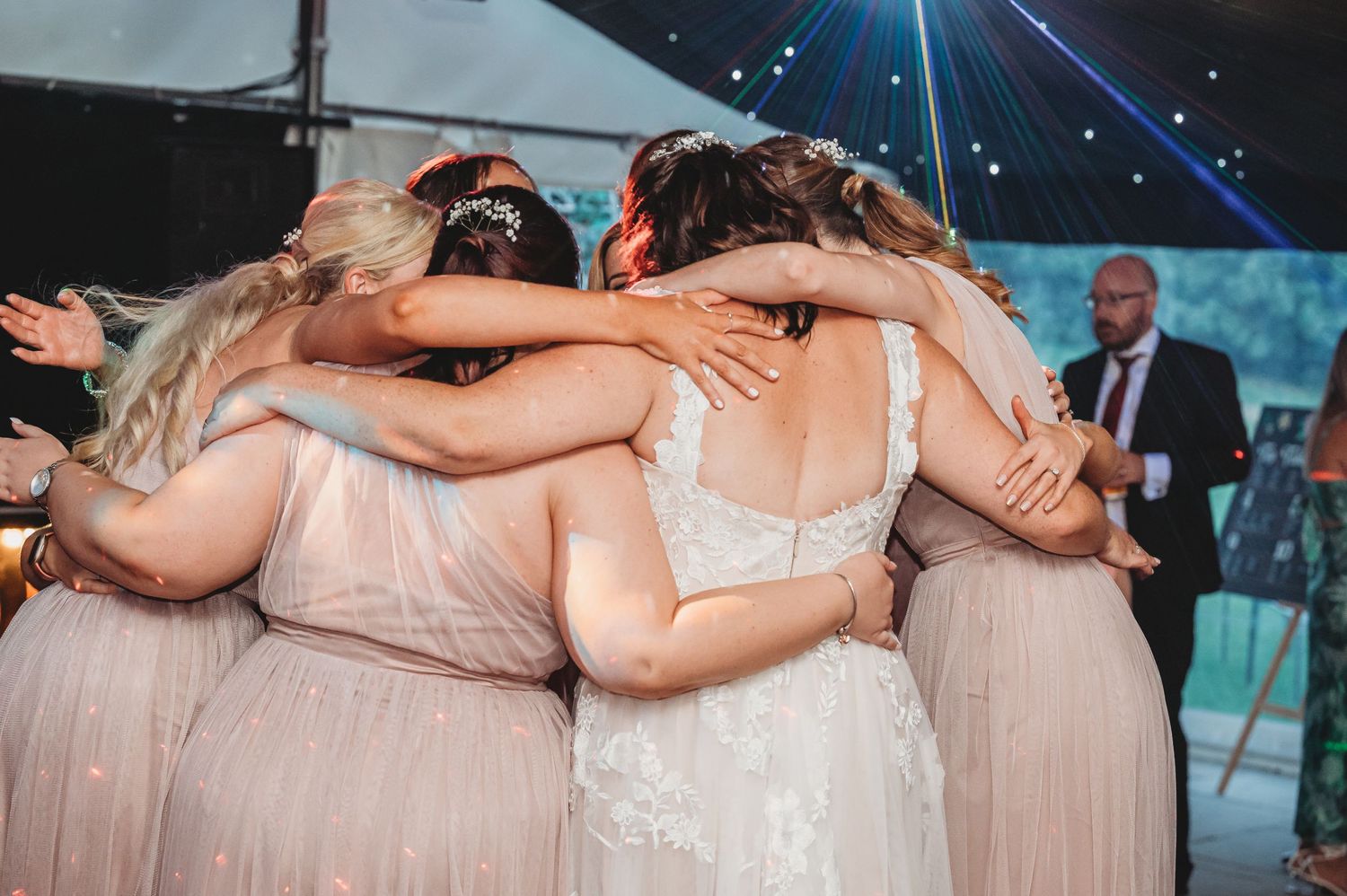 Bridesmaids in blush pink dresses share an emotional group hug during wedding celebration.