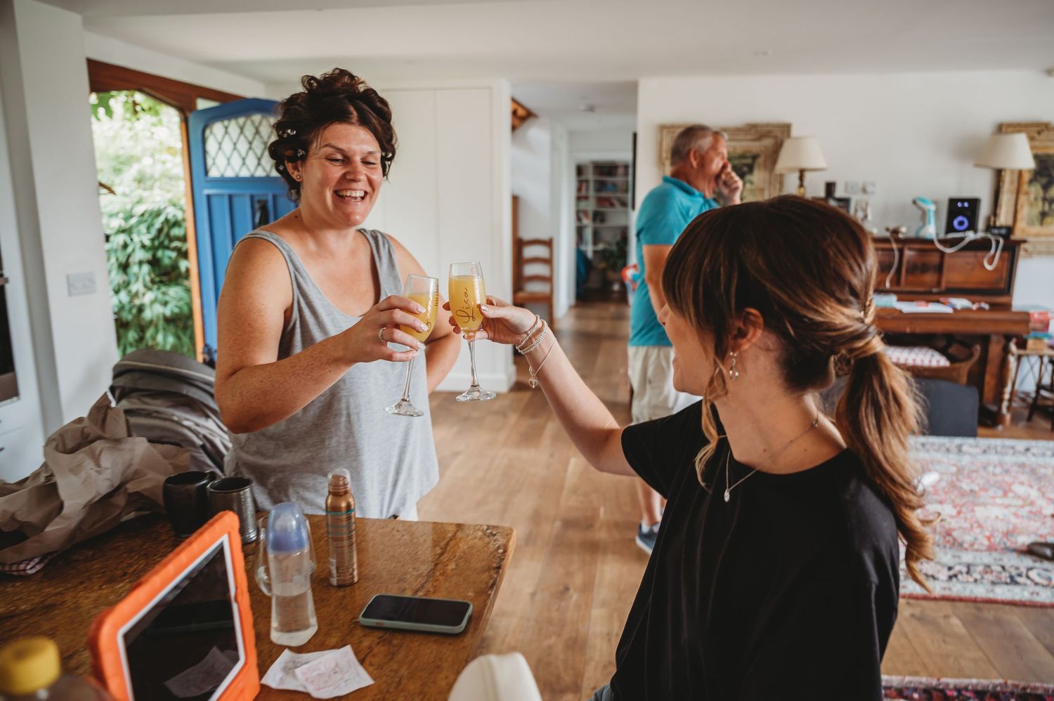 Two people cheerfully toast drinks together in a bright, sunlit kitchen with wooden countertops.