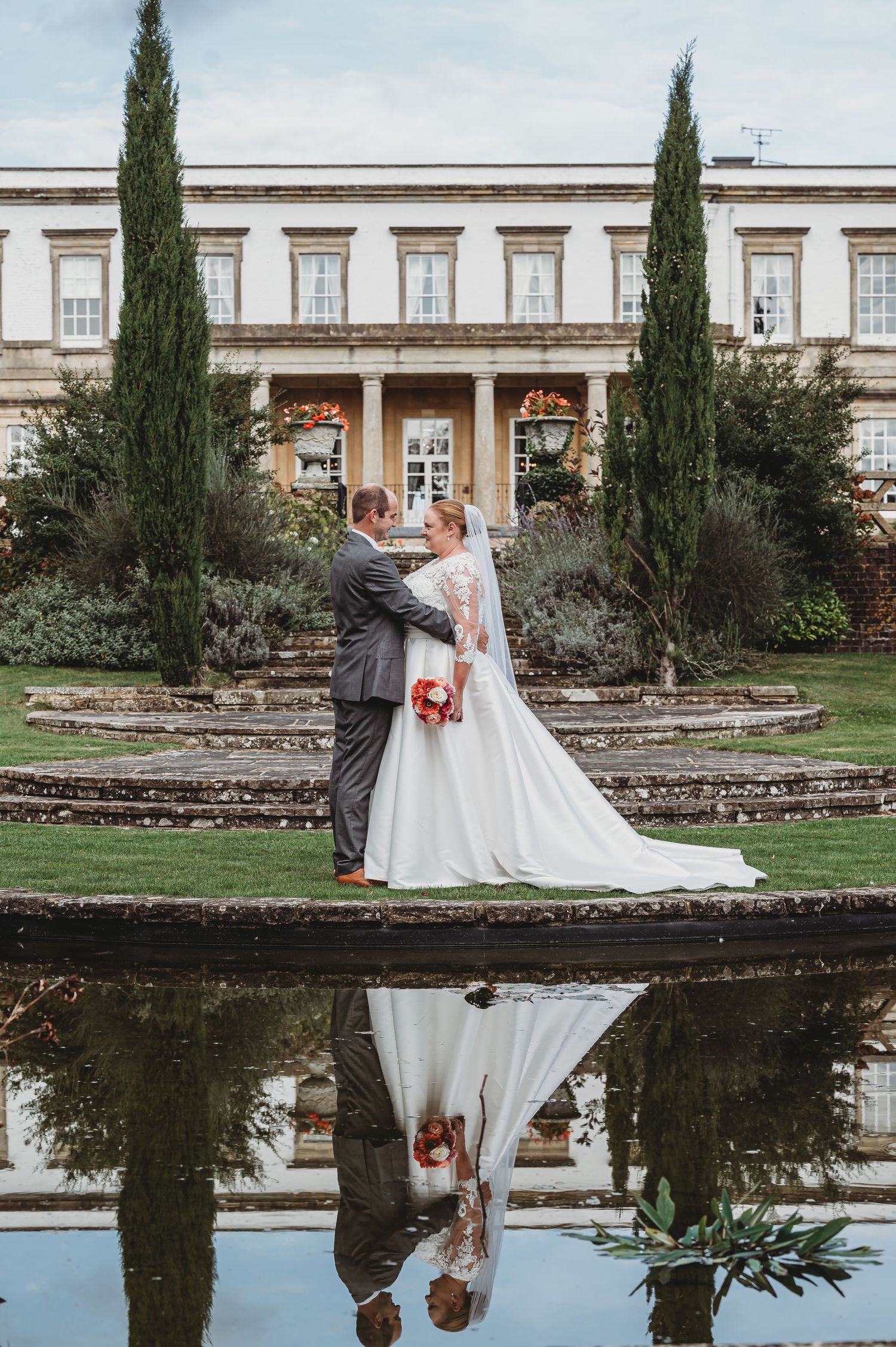 A wedding couple stands between tall cypress trees outside a grand estate with their reflection in a pond below.