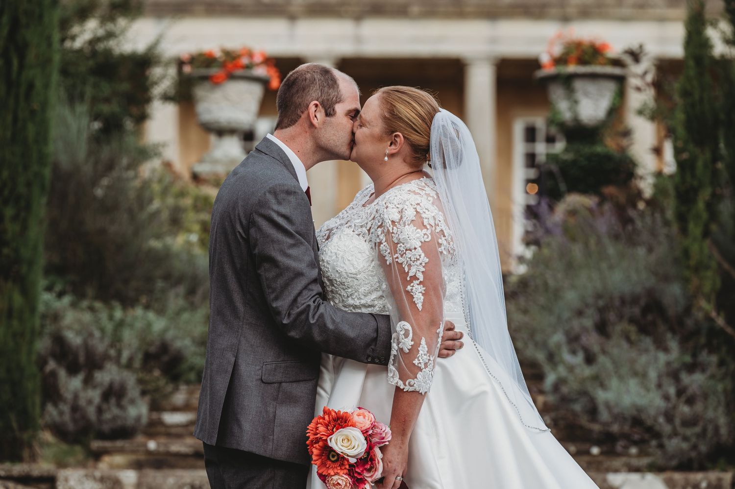 Newlyweds share a romantic kiss in a garden courtyard while holding a vibrant coral bouquet.