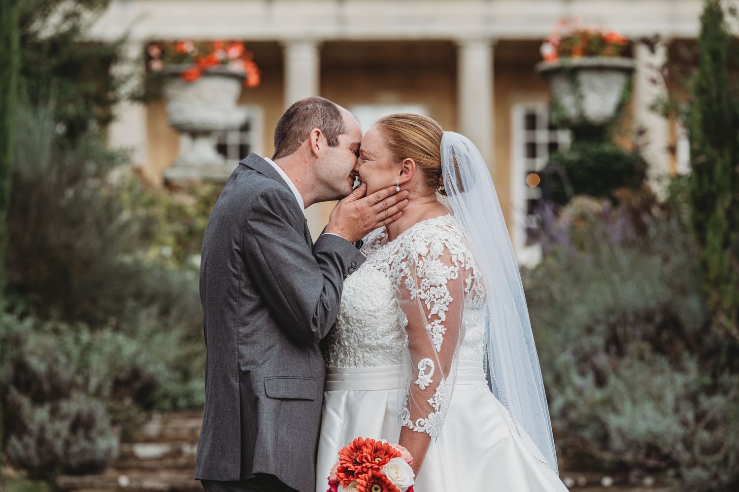A romantic wedding photograph showing a bride in lace and groom in gray suit embracing in a garden setting.