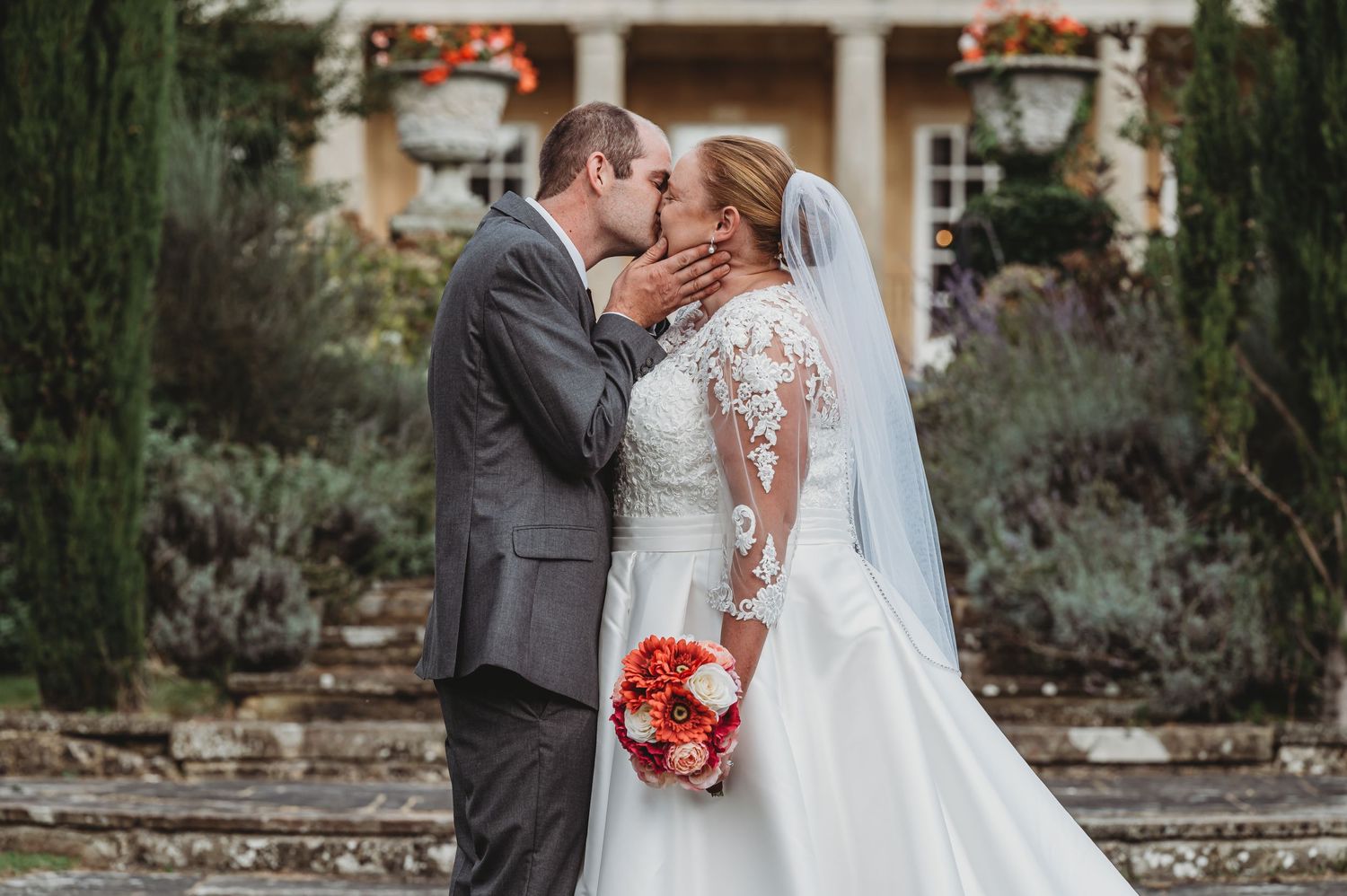 A bride in a white lace gown with a veil kisses their partner in a gray suit while holding a red bouquet.