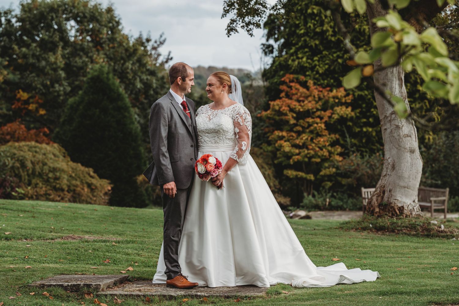 Two people in wedding attire share a romantic moment in a garden setting with autumn foliage in the background.