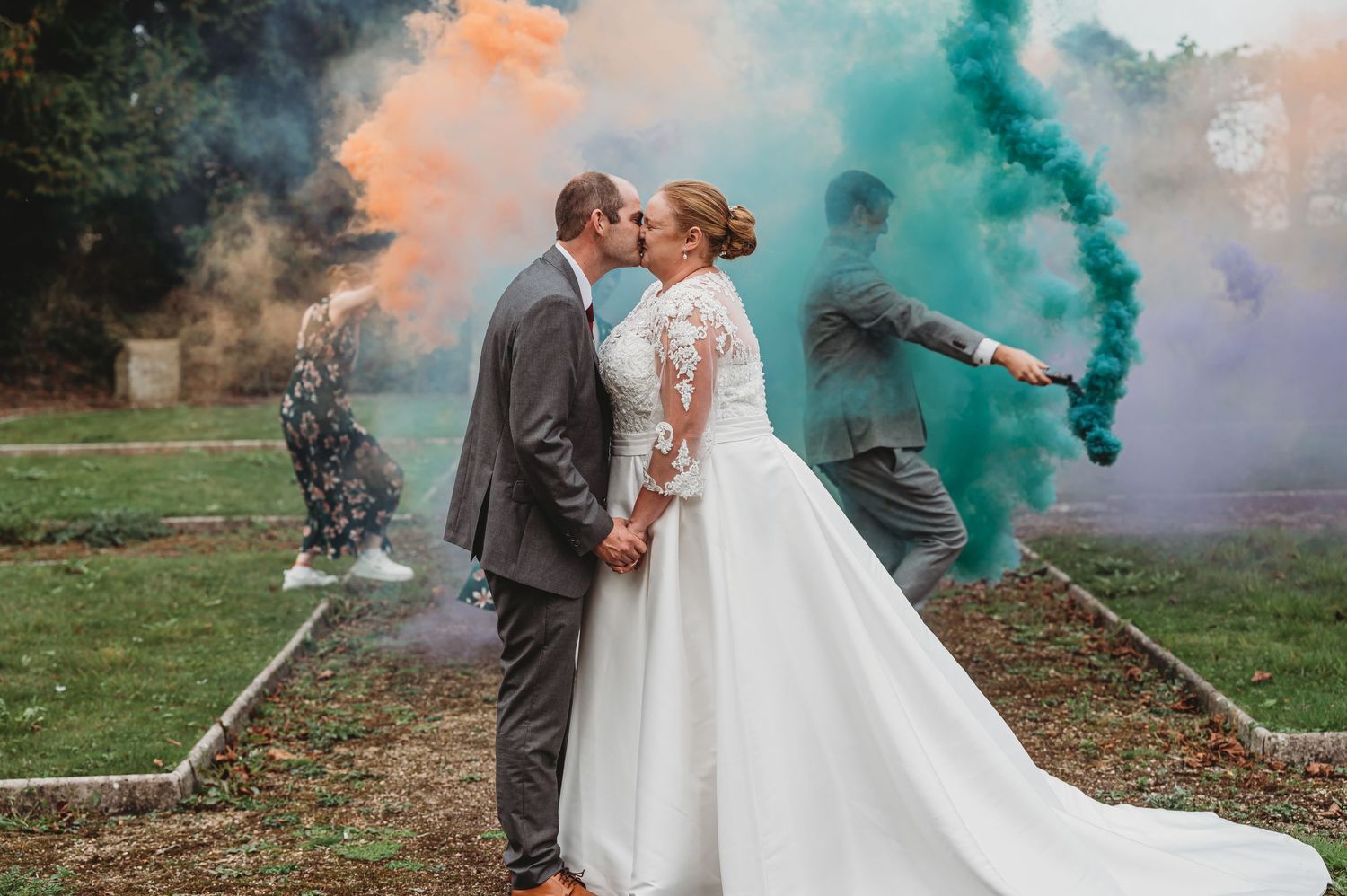 Newlyweds share a romantic moment surrounded by colorful smoke bombs in a garden setting during their wedding photoshoot.