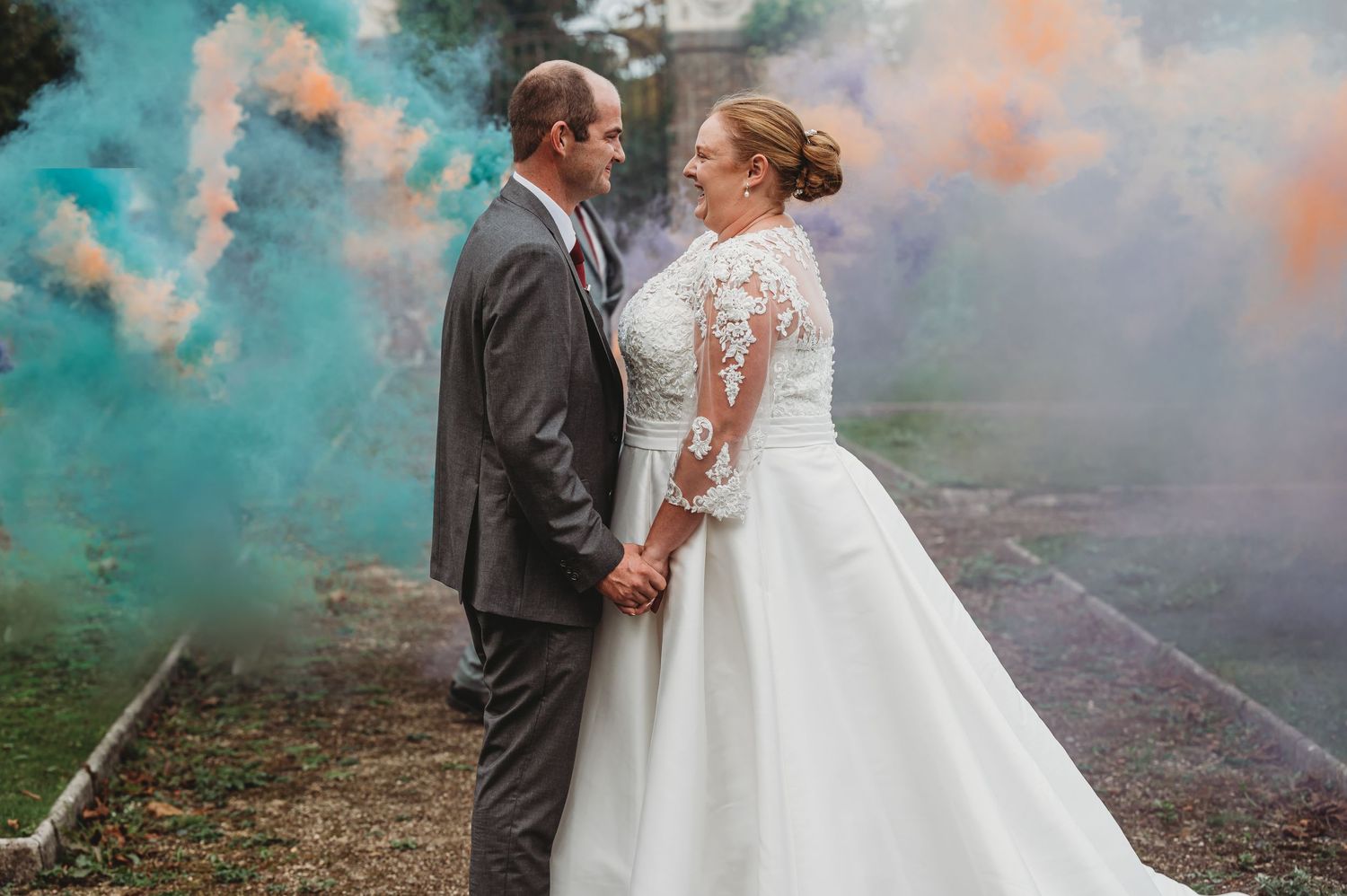 A wedding couple stands together surrounded by colorful teal smoke during their outdoor photography session.