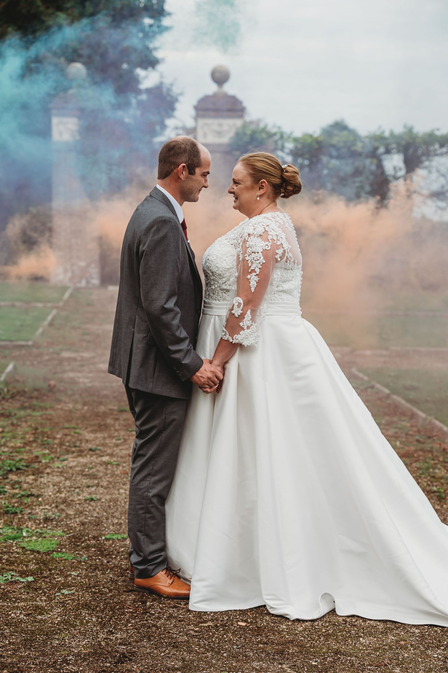A couple in wedding attire holding hands on misty grounds with elegant architectural elements in the background.