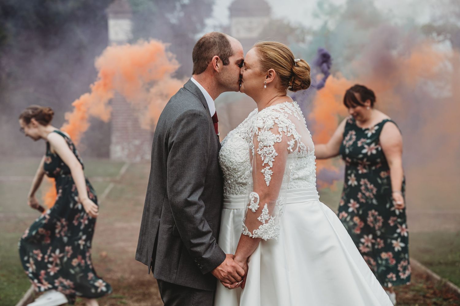 A couple shares a romantic kiss in their wedding attire surrounded by colorful smoke bombs and bridesmaids.
