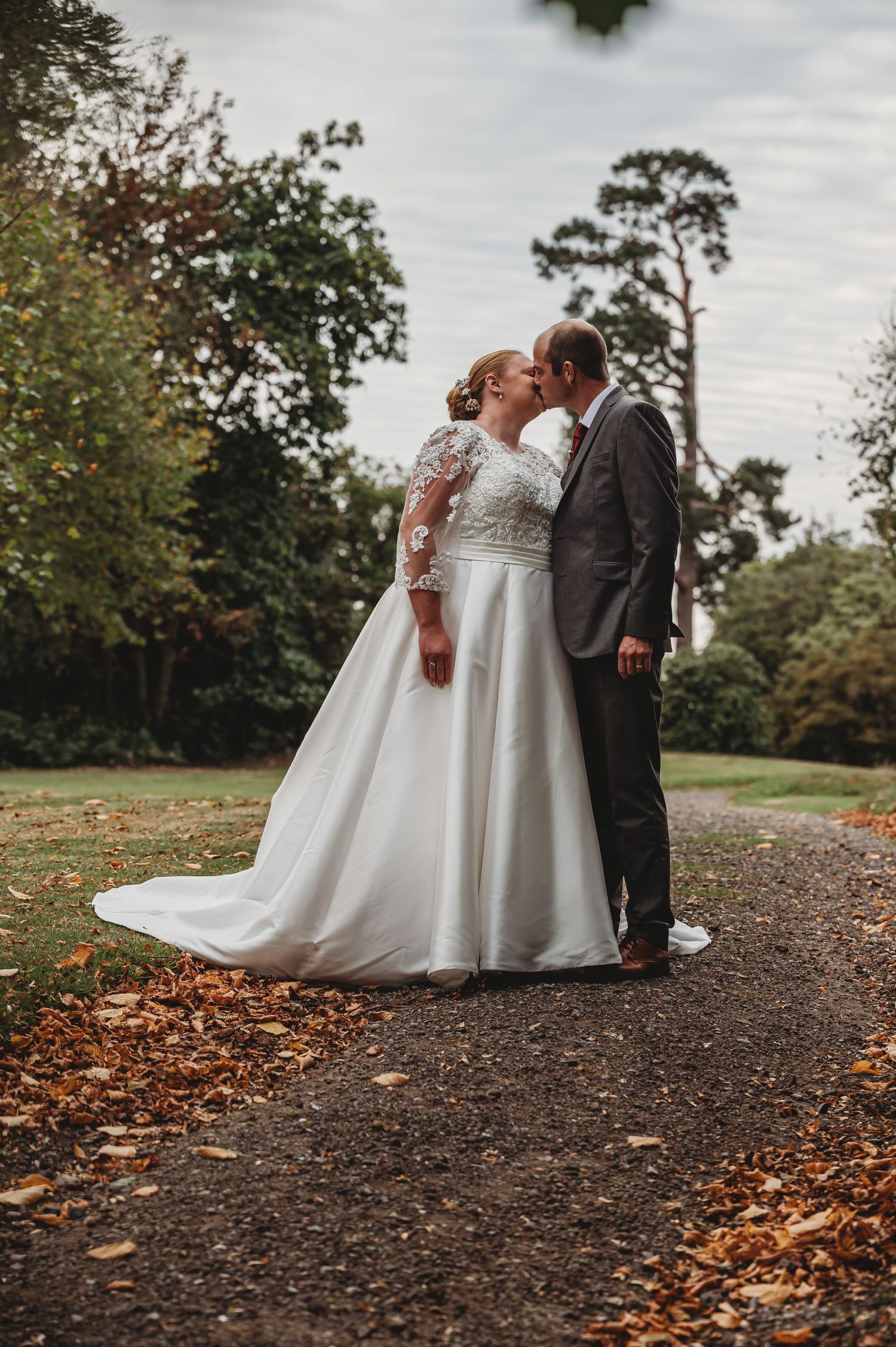 A romantic autumn wedding photo of a couple embracing on a woodland path covered in fallen leaves.