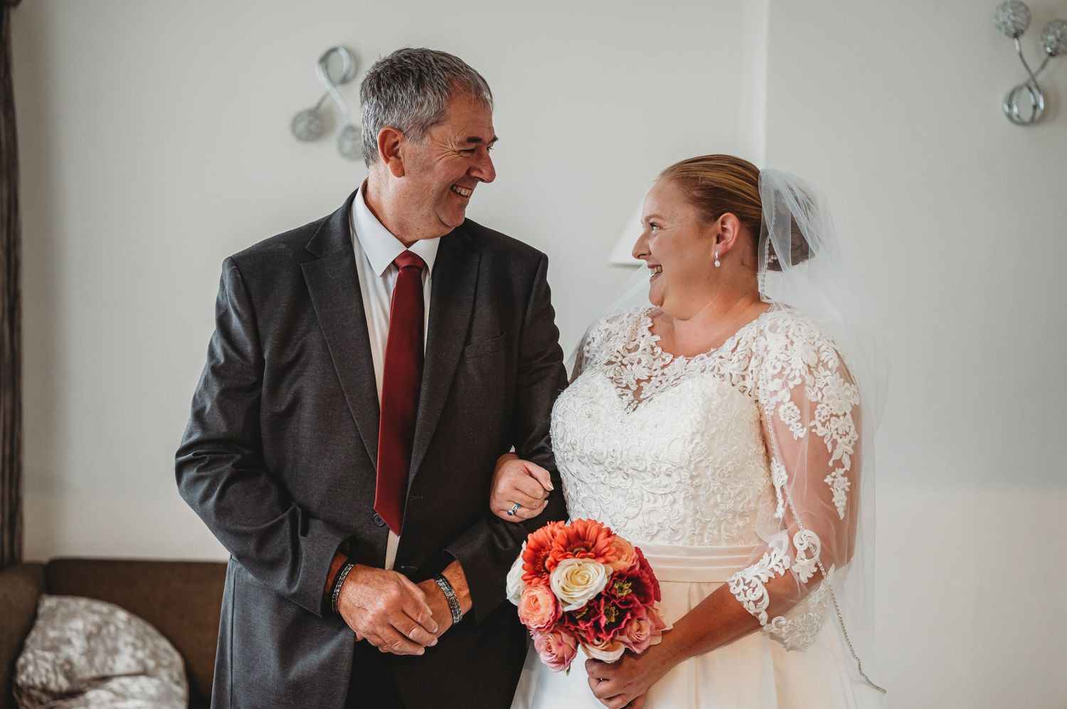 A beaming bride in a white lace dress gazes lovingly at someone in a suit while holding a bright coral bouquet.