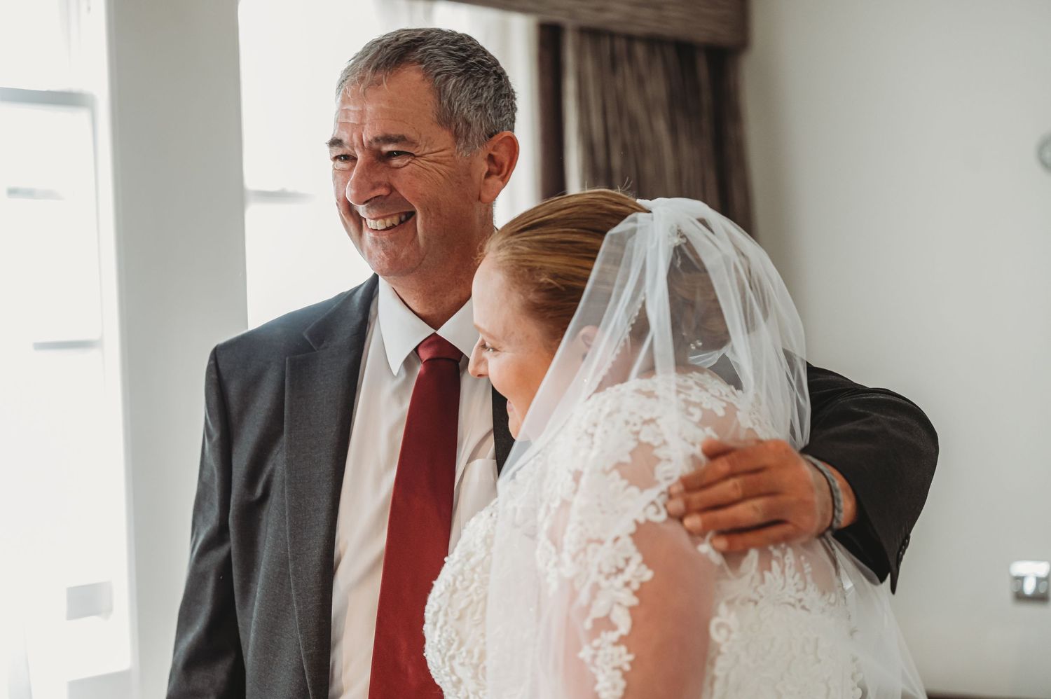 A tender moment between two people at a wedding, with one wearing a white lace veil and another in a suit with red tie.