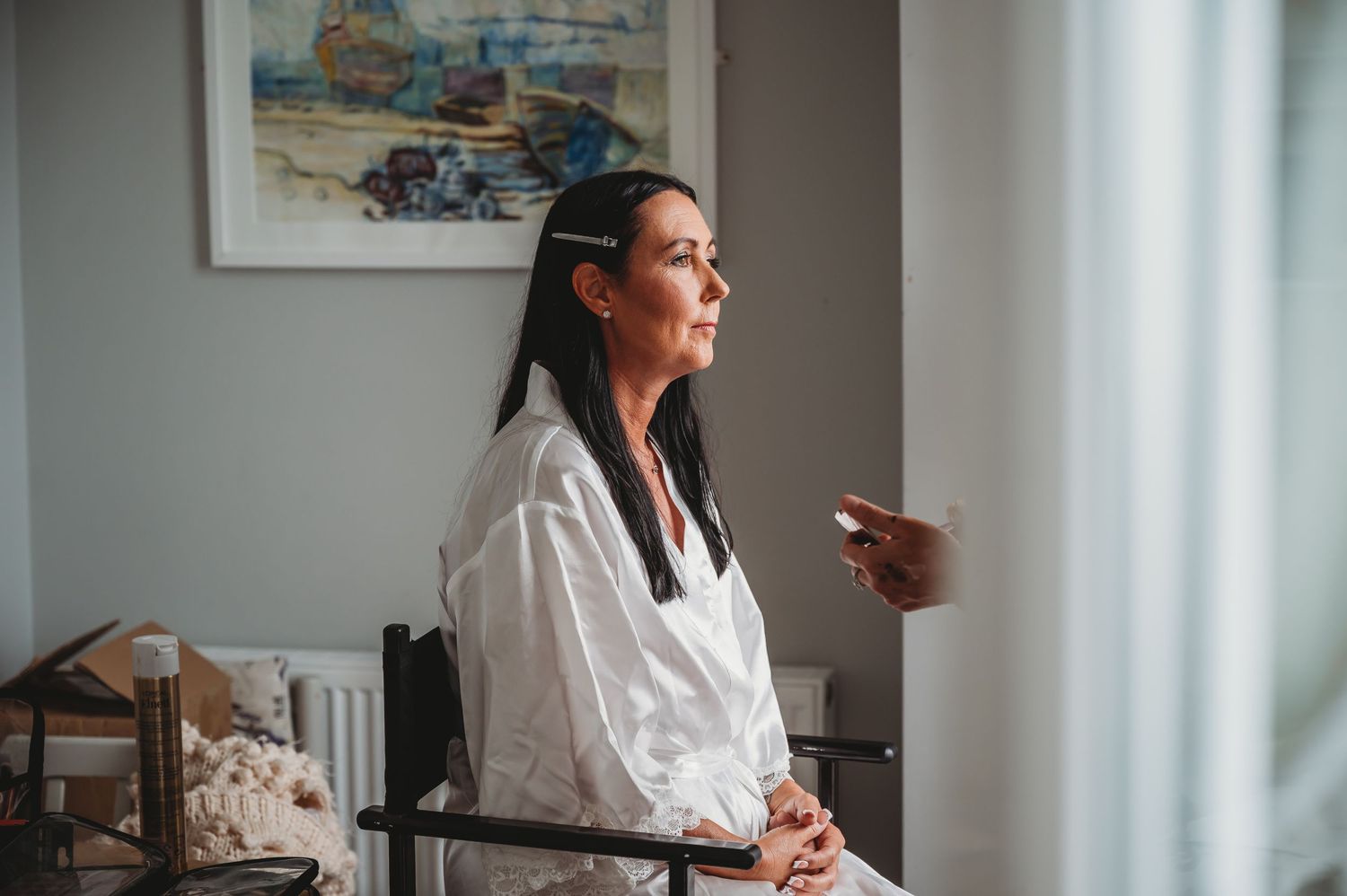 A person wearing a white robe gazes thoughtfully through a window in a room with framed artwork on the wall.