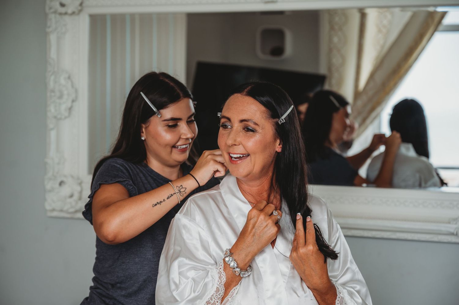 Two people share a joyful moment while getting ready in a bright room with a mirror visible in the background.