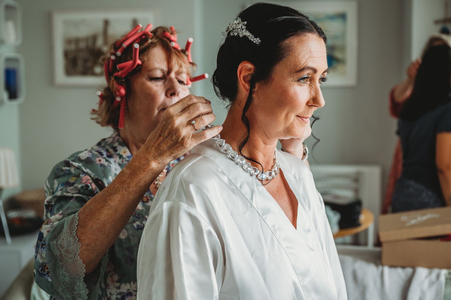 A hairstylist in pink curlers works on a person in white robe during wedding day preparations.