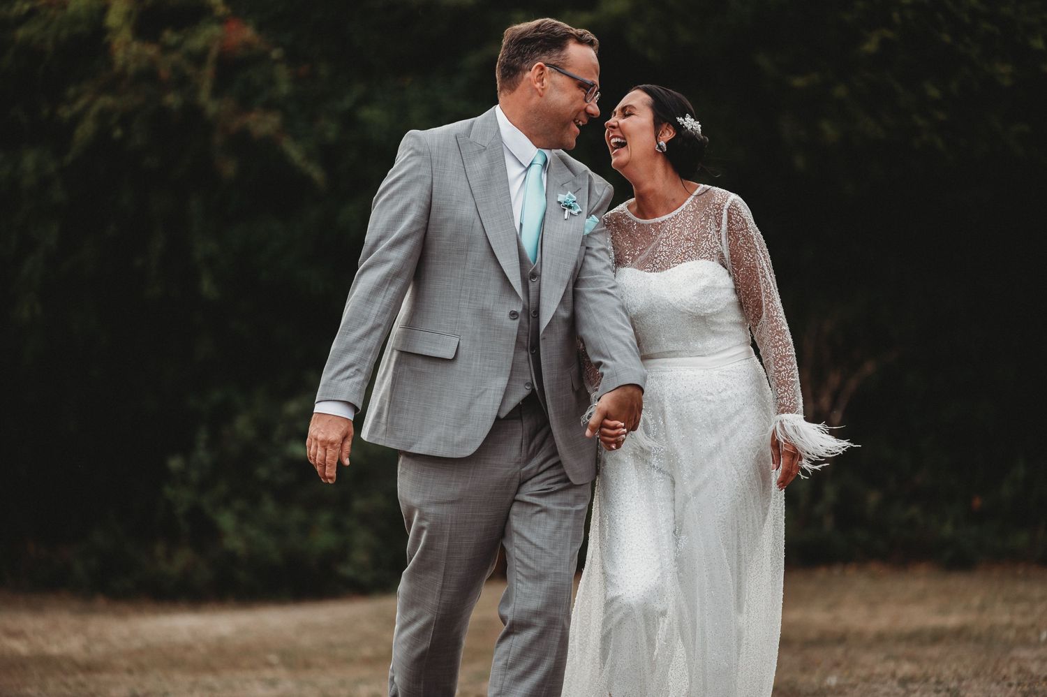 Newlywed couple in gray suit and white lace dress share romantic moment while holding hands outdoors.