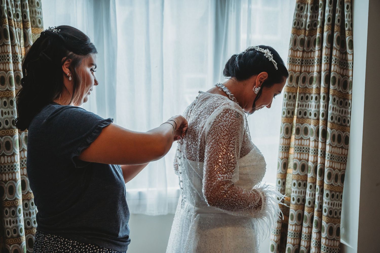 The bride gets help putting on a delicate lace wedding dress near a window with patterned curtains.