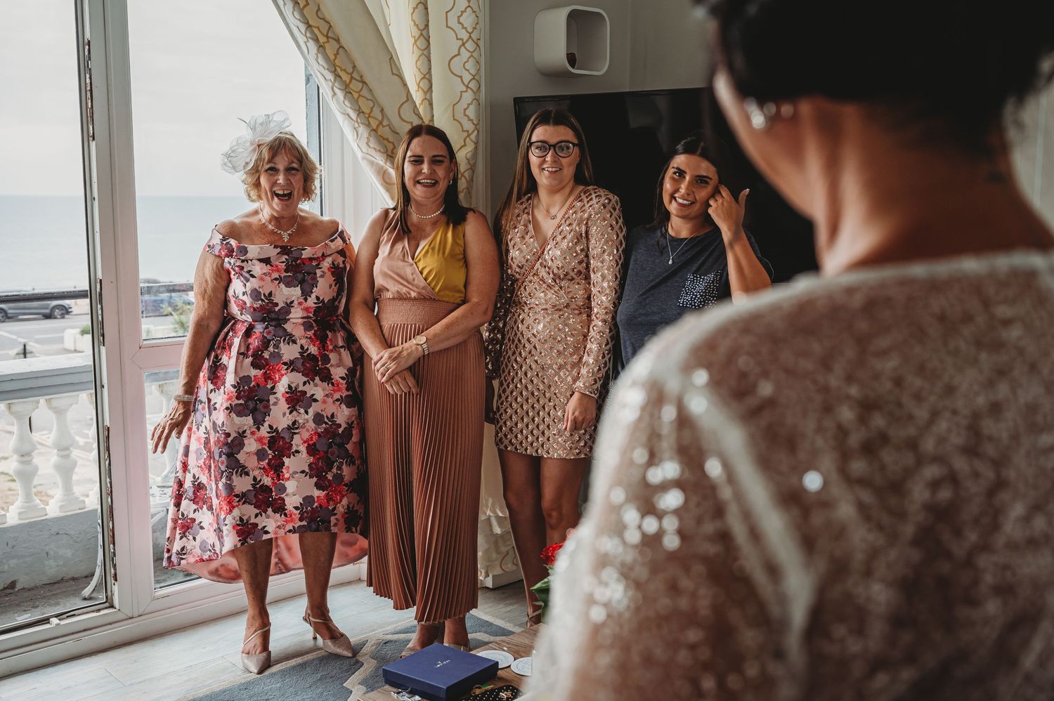 A group of people stand by a bright window wearing elegant dresses in floral prints and solid colors during a special occasion.