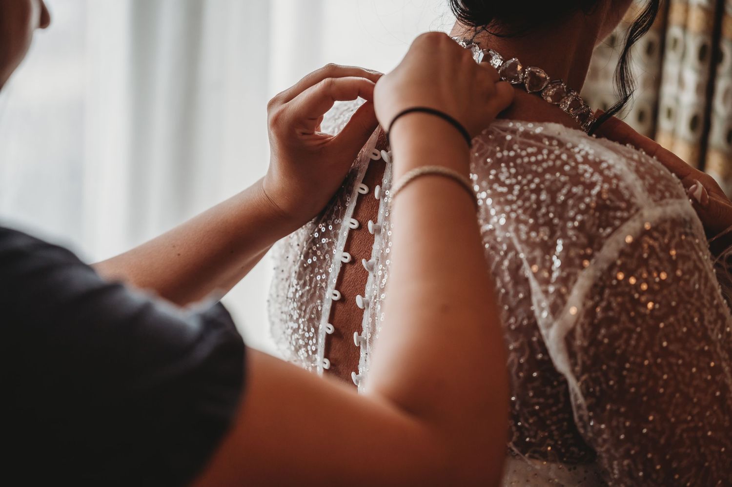 Close up view of buttoning a delicate sheer wedding dress covered in intricate beadwork and sparkles.