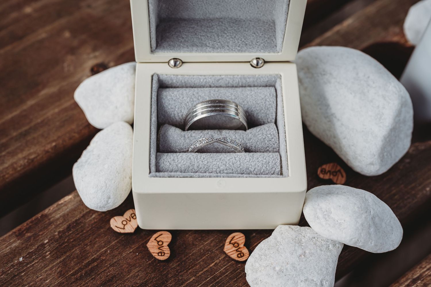 Wedding ring in a white jewelry box surrounded by smooth white stones and decorative pebbles on wooden surface.