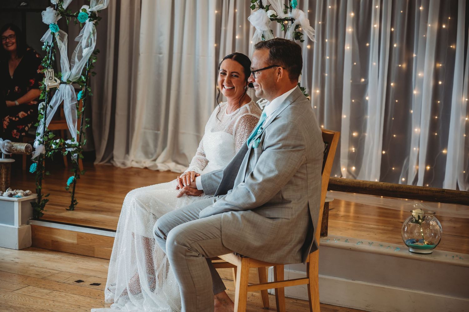 A couple sits together during their wedding ceremony with white curtains and blue decor in the background.