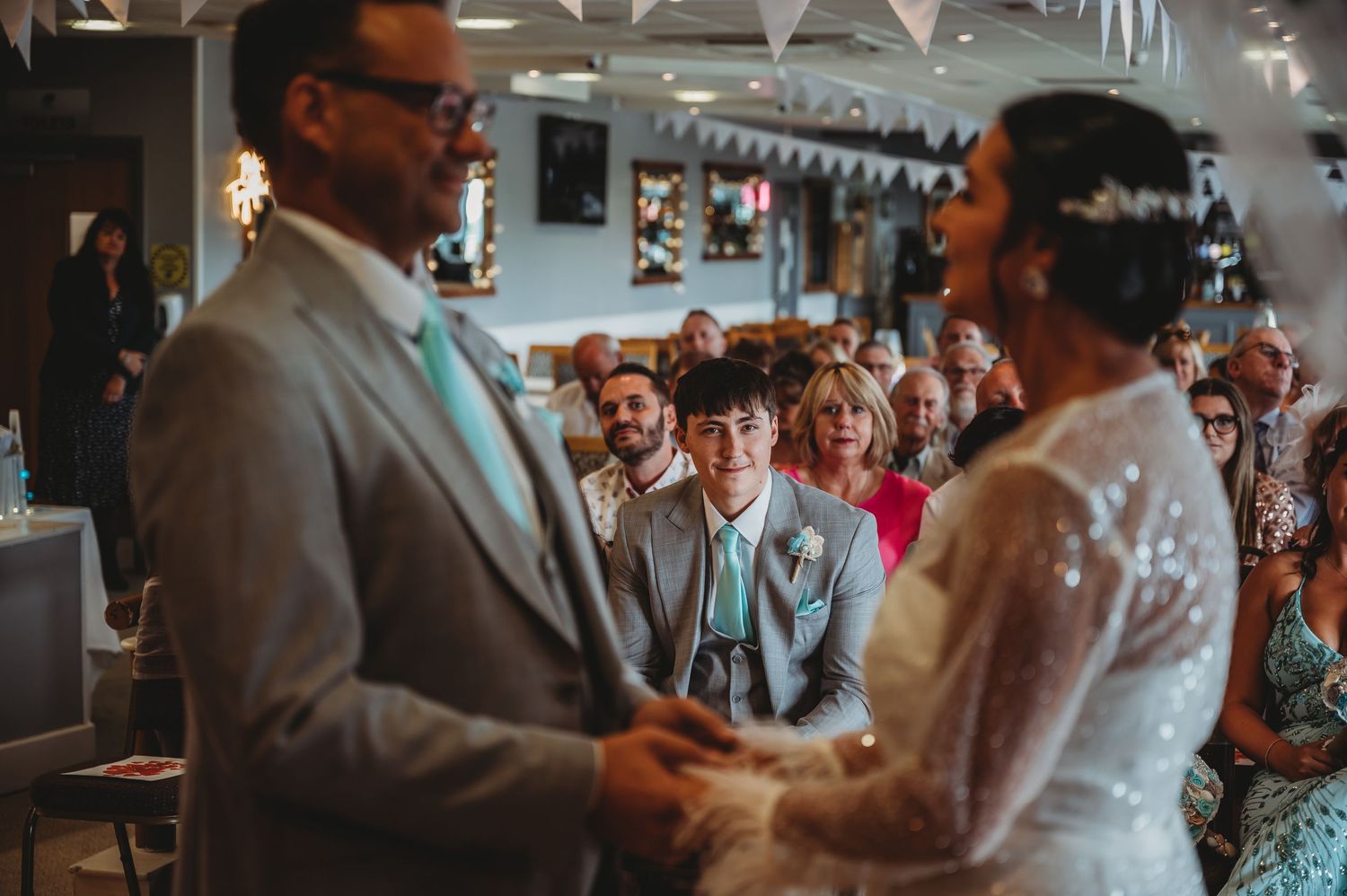 A couple exchanges vows during an indoor wedding ceremony surrounded by guests and decorative bunting.