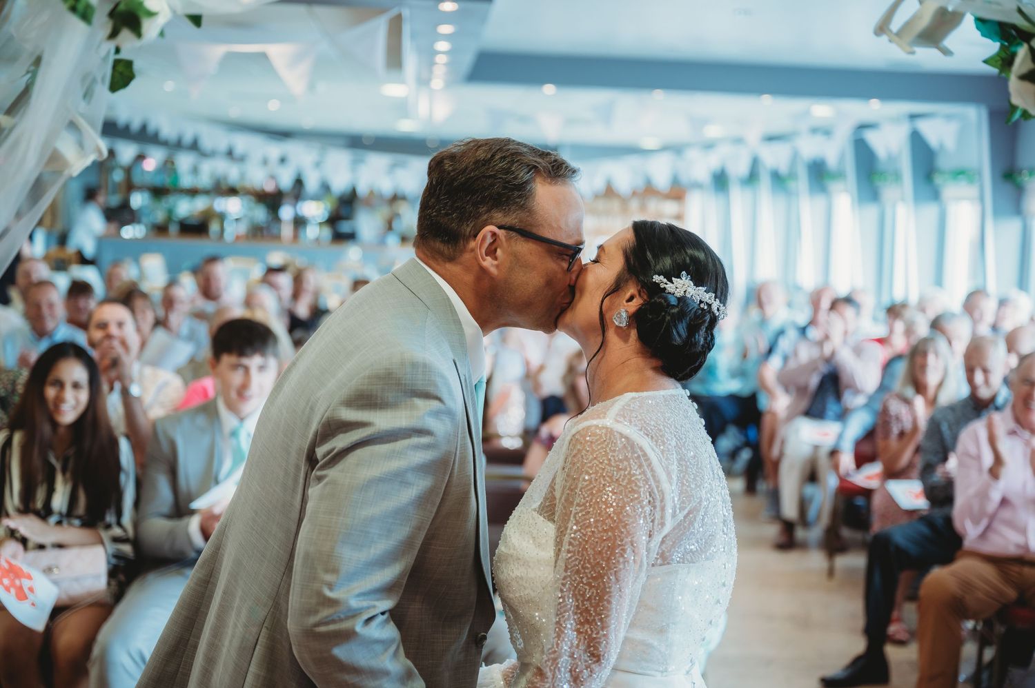 A couple shares their first kiss at a wedding ceremony while guests look on in a bright, decorated venue.