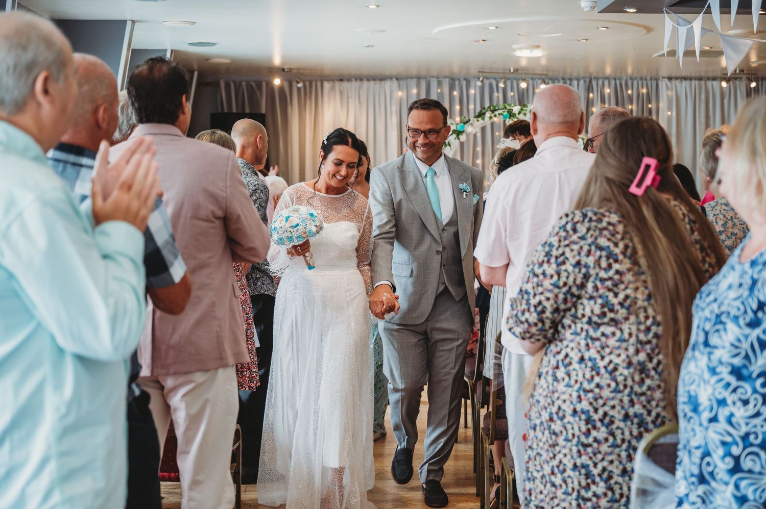 Wedding guests clap and celebrate as newlyweds in white dress and gray suit dance and smile during indoor reception.