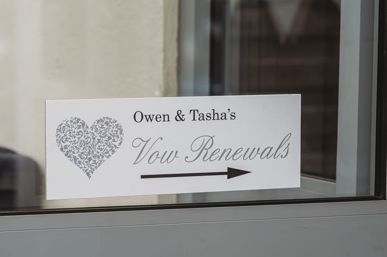 A white wedding sign displaying 'Vow Renewals' with a decorative heart design in gray tones sits on a windowsill.
