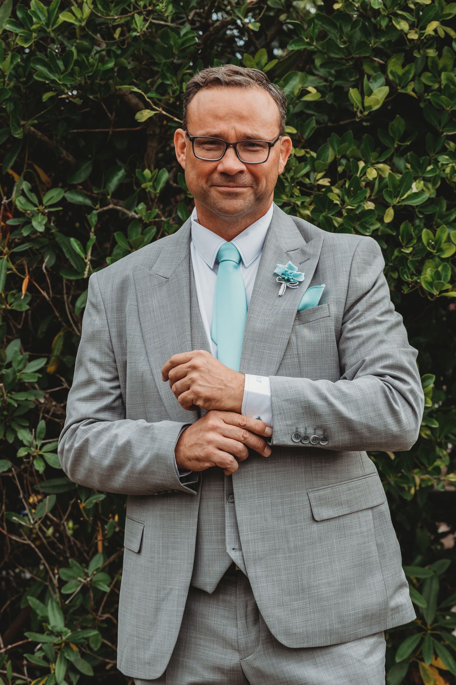 A groom in a light gray suit with mint tie adjusts his cufflink while standing against a green hedge background.