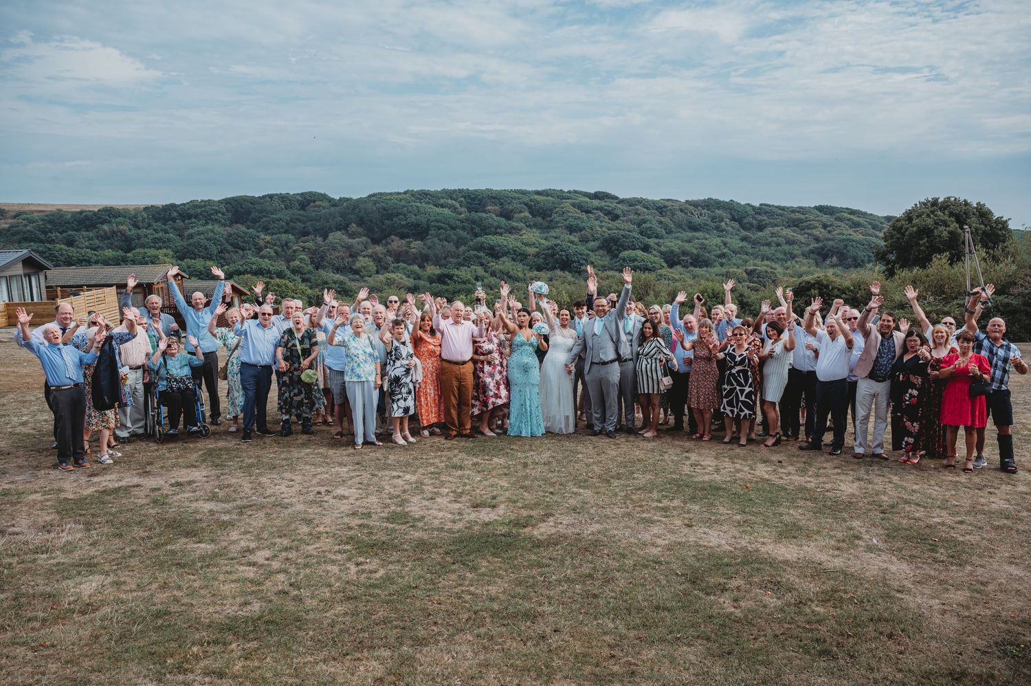 Large group of wedding guests gathered outdoors on a hilltop against a cloudy summer sky landscape.
