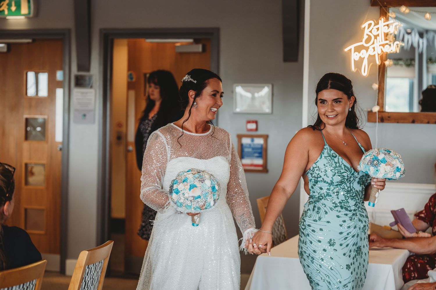Two people in special occasion dresses stand together holding hands, one in white lace and one in sparkly turquoise.