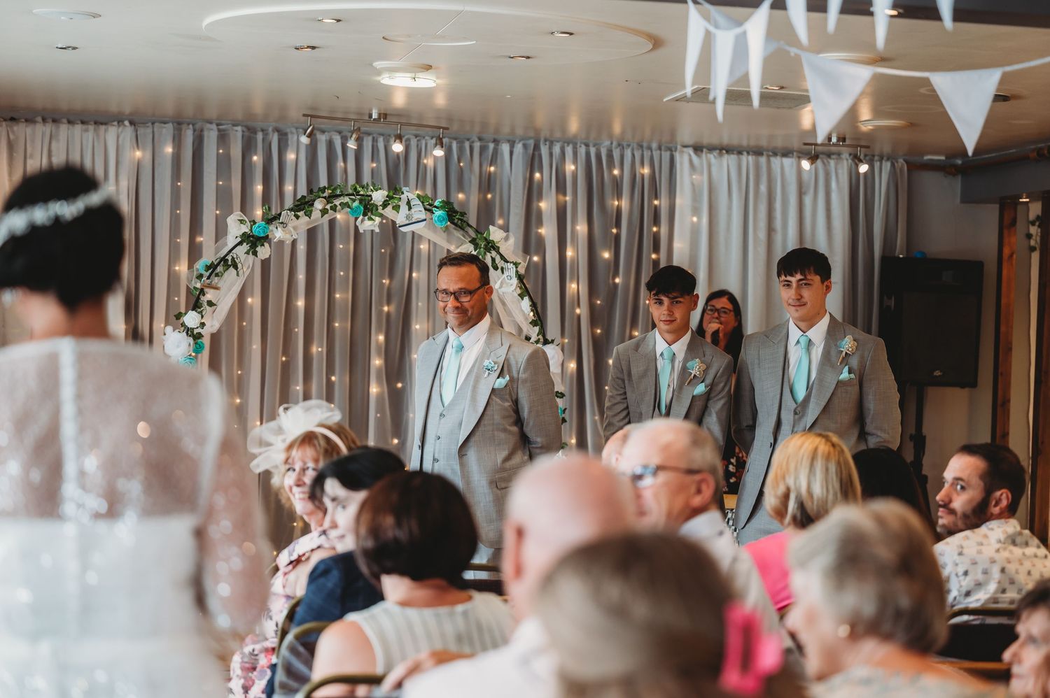 Wedding ceremony taking place under a floral arch with guests seated in a decorated indoor venue.