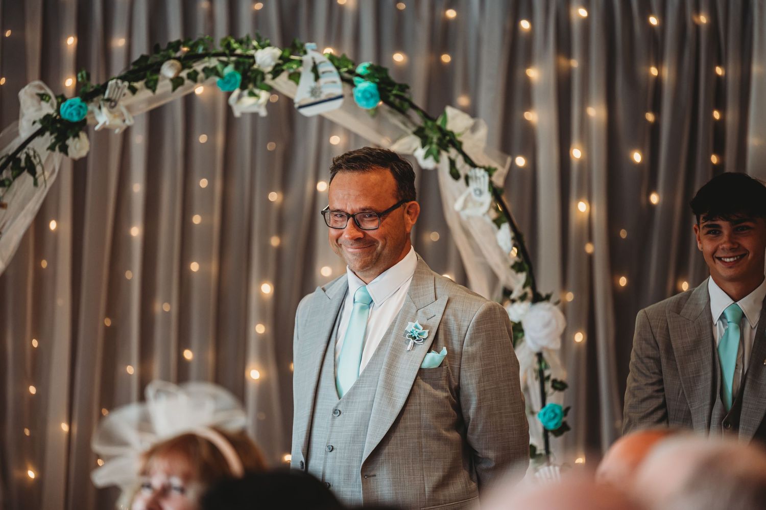 Wedding ceremony setup with a floral arch decorated with teal accents and twinkling lights.