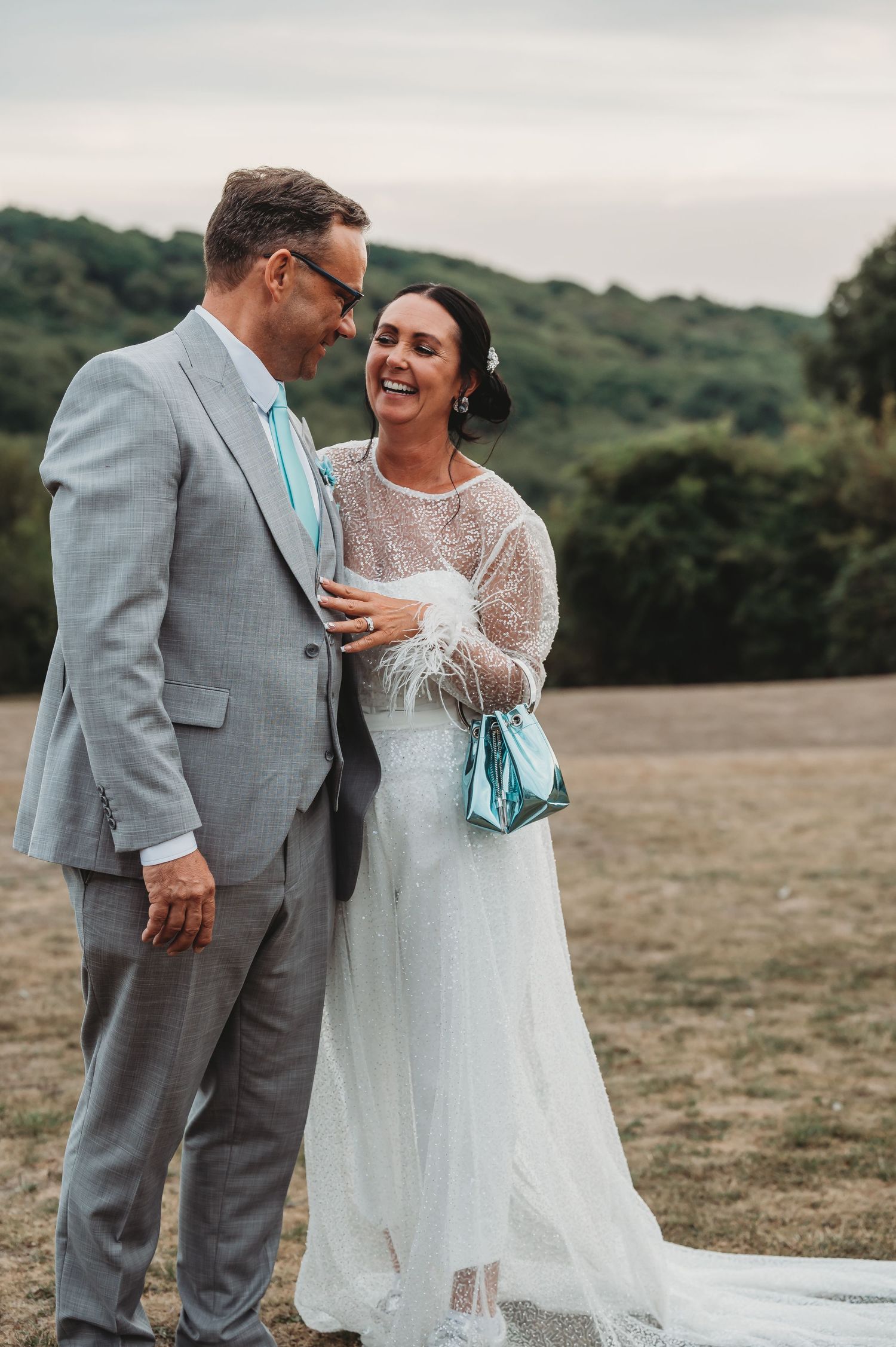 A happy couple in wedding attire laugh together on a hilltop with green hills behind them at sunset.