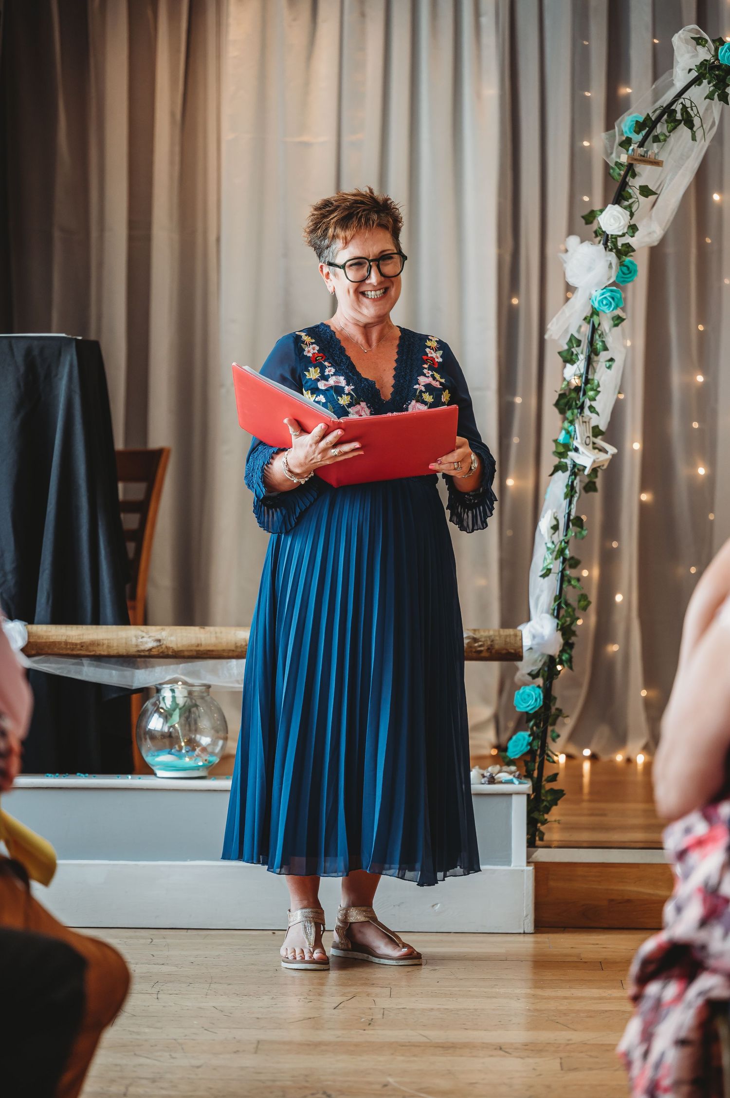 Wedding officiant in navy blue pleated dress holding a red folder stands at a decorated ceremony venue with twinkle lights.