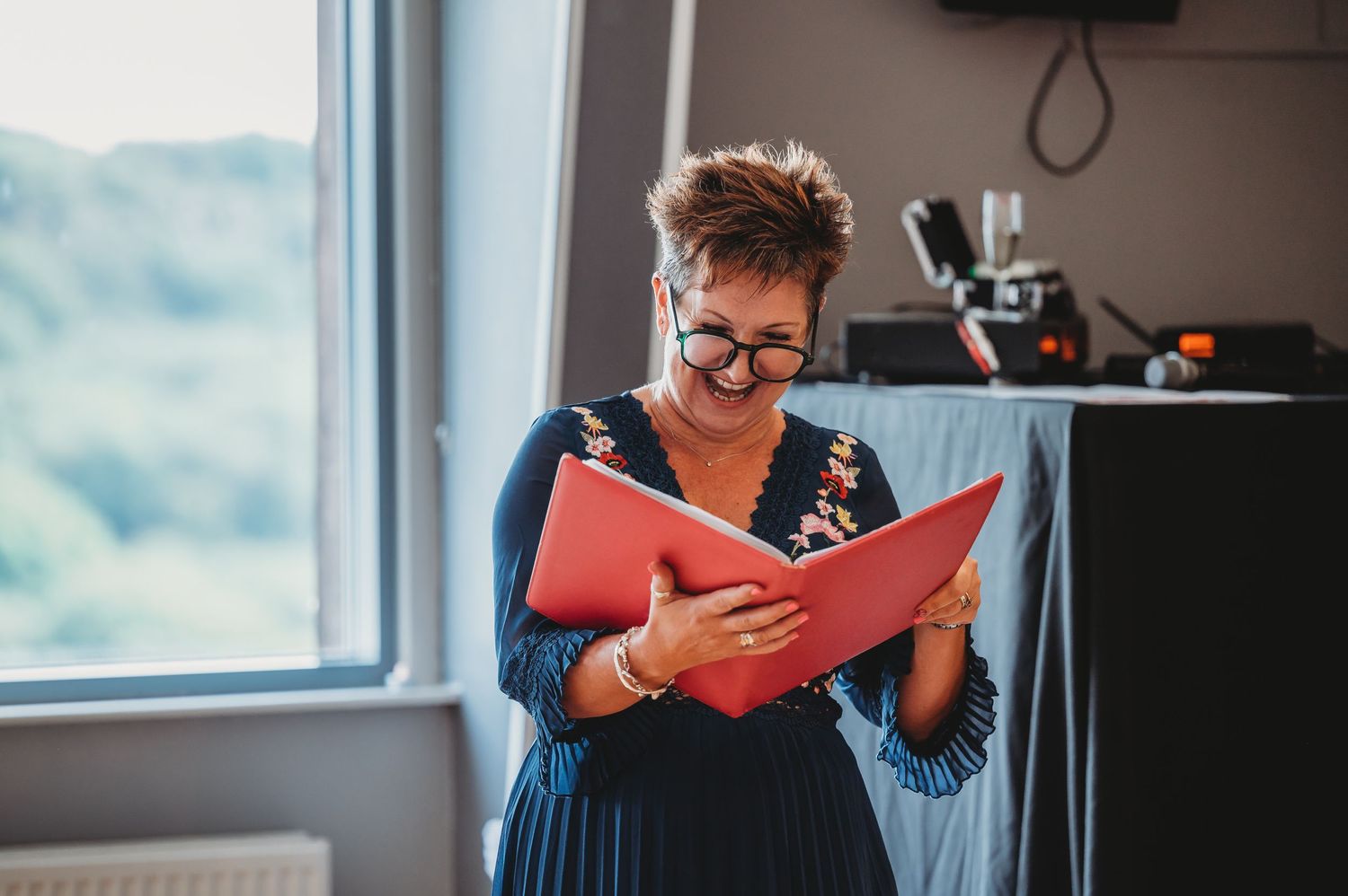 Someone in navy dress and glasses reads from a red folder while standing by a window in natural light.