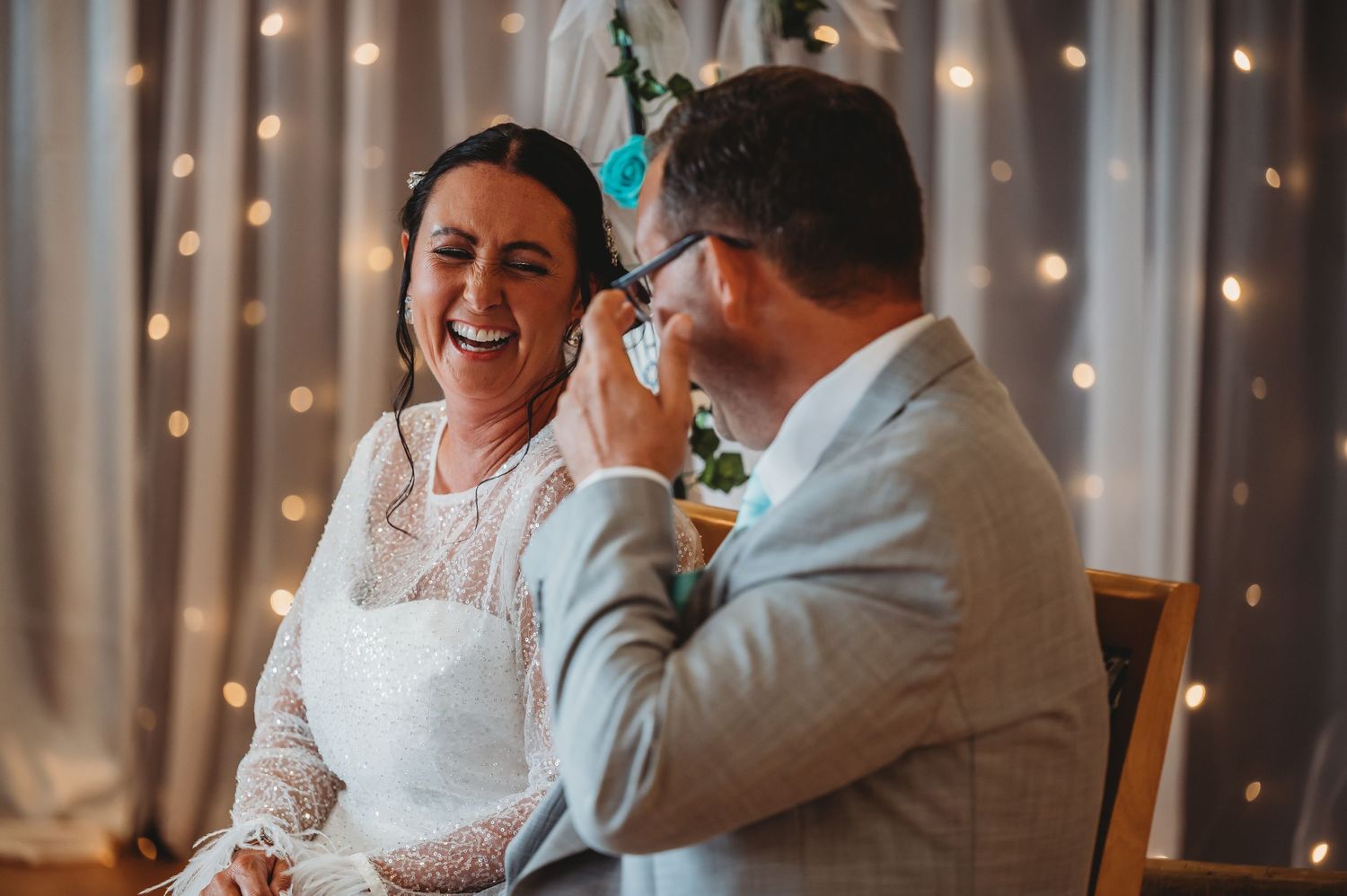 Couple shares a joyful candid moment at wedding reception with twinkle lights glowing in background.