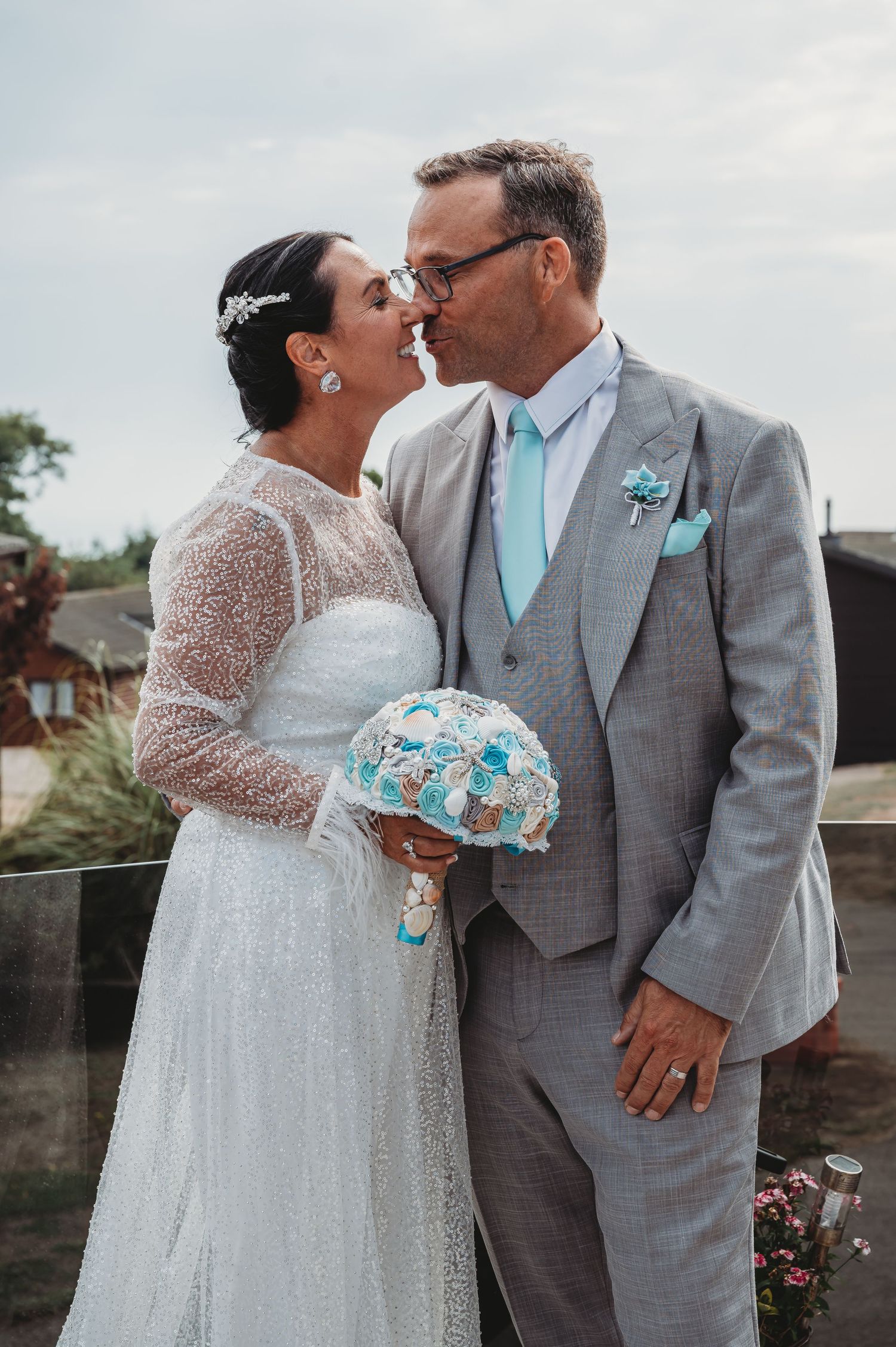 A happy wedding couple shares a tender moment outdoors, with the bride holding a blue and white bouquet.