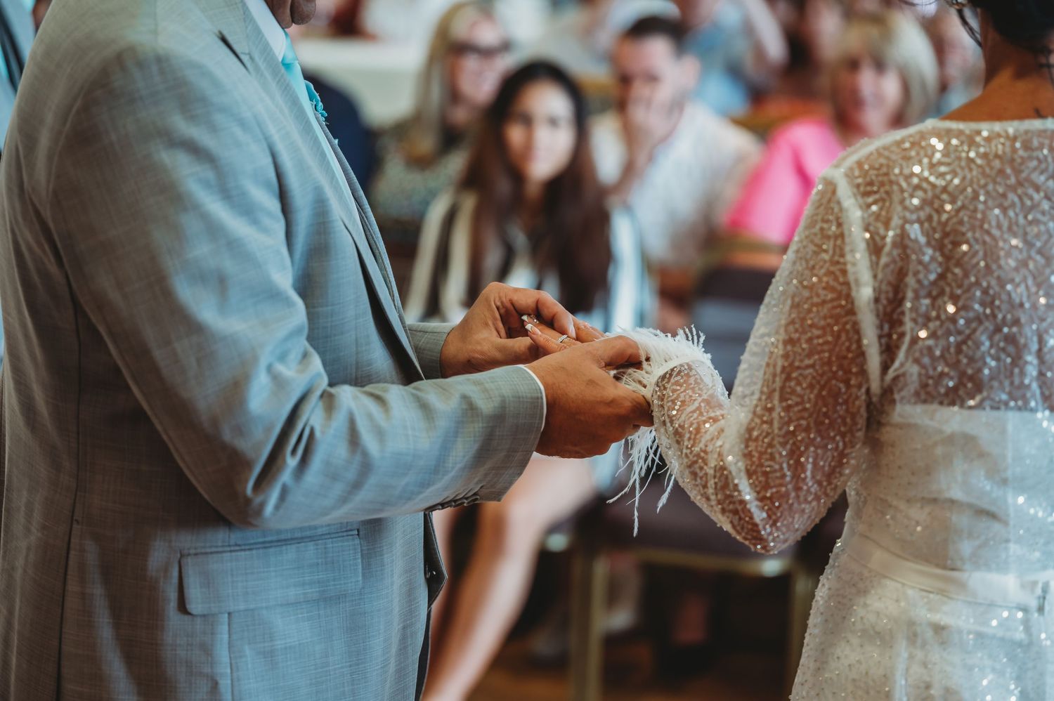 Close-up view of wedding ring exchange during ceremony with suit sleeve and lace dress details in soft focus.