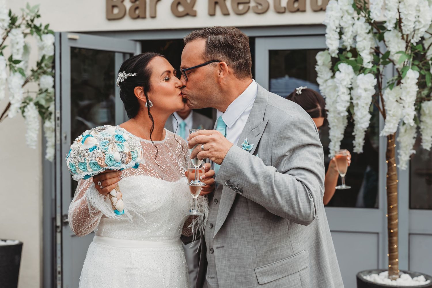 Newlyweds share a romantic kiss while holding champagne glasses at an outdoor bar decorated with floral archway.