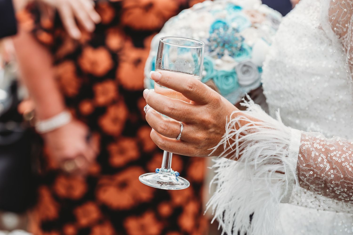 A hand wearing a white lace sleeve holds a blue cocktail in a wine glass against an orange floral background.